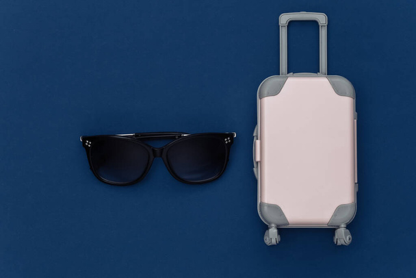Die Reise lag flach. Mini-Reisekoffer aus Kunststoff, Sonnenbrille auf klassisch blauem Hintergrund. Minimaler Stil. Ansicht von oben - Foto, Bild