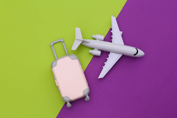 航空旅行フラットレイアウト。緑の紫色の背景にミニプラスチック旅行スーツケースと航空機。トップ表示. - 写真・画像