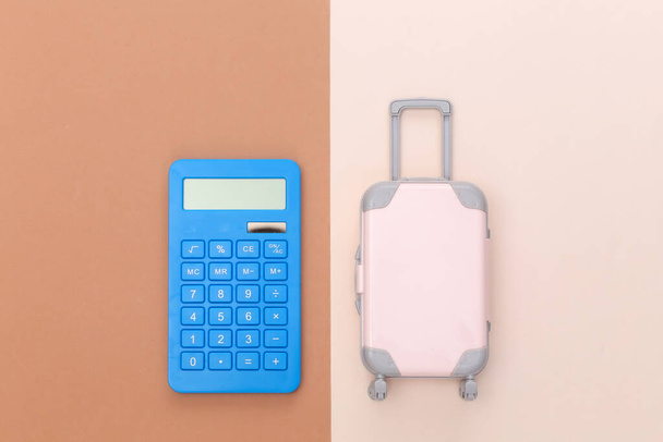 Путевые расходы. Мини-пластиковый дорожный чемодан с калькулятором на бежевом коричневом фоне. Вид сверху. Плоский лежал - Фото, изображение