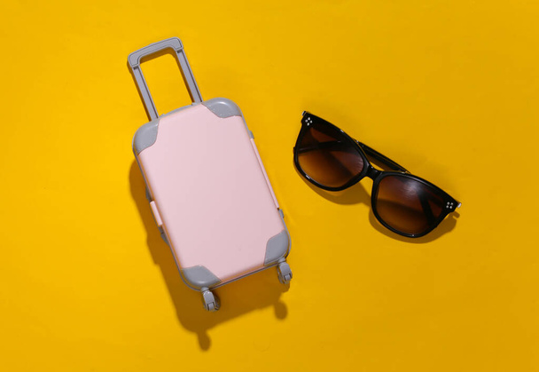 Reisminimalisme. Mini plastic reiskoffer en zonnebril op gele achtergrond met diepe schaduw. Minimale stijl. Bovenaanzicht, vlak lay - Foto, afbeelding