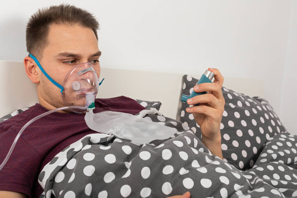 Male patient with oxygen mask at home is lying in bed, holding an inhaler spray with salbutamol - Foto, Bild