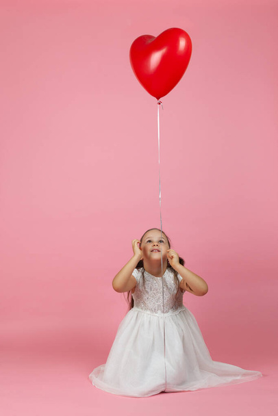 Ένα άτακτο, παιχνιδιάρικο παιδί με αλογοουρές και λευκό φόρεμα κάθεται στο πάτωμα και κοιτάζει ένα κόκκινο μπαλόνι σε σχήμα καρδιάς από πάνω της, απομονωμένο σε ροζ φόντο. - Φωτογραφία, εικόνα