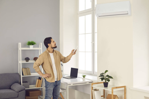 Man standing in living-room and switching on air conditioner with remote control - Foto, afbeelding