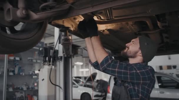 Prise de vue à la main du mécanicien automobile masculin barbu en salopette et gants faisant des réparations sur le train d'atterrissage de la voiture dans le garage - Séquence, vidéo