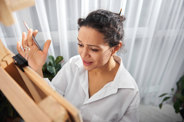 A young artist stands in front of an easel and nervously spreads her hands holding a pencil - Photo, Image