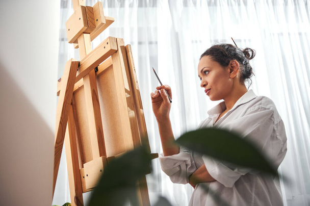 Beautiful woman painter standing in front of the easel in workshop - Foto, afbeelding