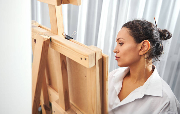 Πλάγια άποψη ενός όμορφου μελαχρινού ζωγράφου με μαζεμένα μαλλιά σε ένα κουλούρι και πινέλα μπογιάς στα μαλλιά της στέκεται μπροστά από το καβαλέτο και ζωγραφίζει - Φωτογραφία, εικόνα