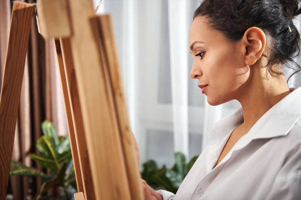 Side portrait of a beautiful woman painter wearing a white shirt and standing in a front of easel. - Фото, зображення