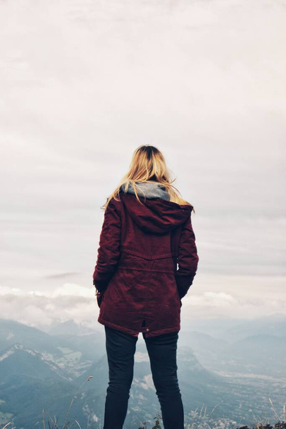 Young blond woman facing away standing on a cliff in winter wearing dark red burgundy coat looking into the distance to the foggy mountains - Photo, Image
