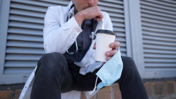 コーヒーと保護医療マスクのカップで手のクローズアップ。疲れ、朝のコーヒーブレーク屋外病院中に医療白衣の疲れ医師。COVID-19, coronavirusパンデミック. - 写真・画像
