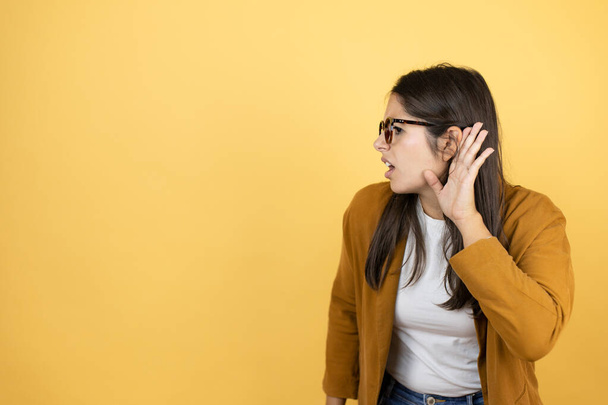 Молодая красивая женщина в блейзере на изолированном желтом фоне с рукой над ухом слушает слухи или сплетни - Фото, изображение