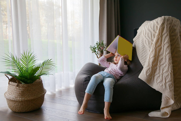 Little cute girl sitting on a bean bag with a book, looking at the window. She's bored - Fotografie, Obrázek