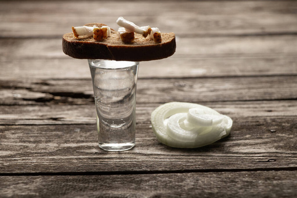 склянку горілки біля шматка чорного хліба з беконом на дерев'яному столі на кухні на обід українською. - Фото, зображення