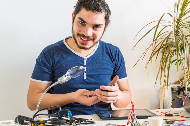 A young repairman of phones and tablets, smiles at the camera, during his work at home. - Fotó, kép
