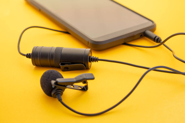 Lavalier or lapel microphone on a colored surface plugged into a cell phone. Details of the grip clip or fastener, mic power bank, miniplug, and wind sponge are visible. Concept useful object, sound production, videobloging. - Photo, Image