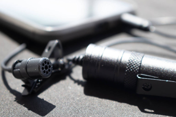 Lavalier or lapel microphone on a black surface, very close-up. The details of the grip clip or bra, , powerbank and conection to cellphone. - Photo, Image