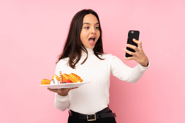 Young brunette woman holding waffles over isolated pink background holding coffee to take away and a mobile - Photo, Image