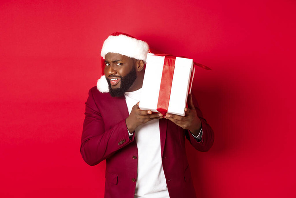 Navidad. Curioso hombre negro en sombrero de santa sacudiendo regalo de año nuevo, preguntarse qué dentro de la caja, de pie sobre fondo rojo - Foto, imagen