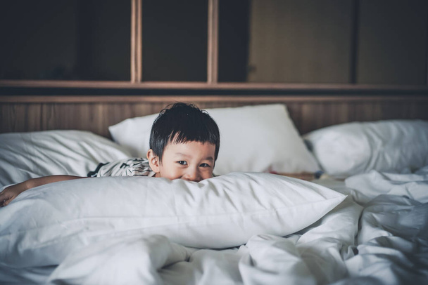 2 jaar oude kleine jongen gekleed in gestreepte T-shirt zijn ontspannen en liggen in bed, warme en gezellige bed vibes scene. Portret van een mooie jongen kijkt naar de camera en voelt zich levend en glimlachend. - Foto, afbeelding