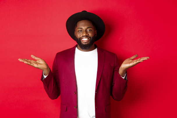 Weihnachtseinkäufe und Menschen-Konzept. Schöner afrikanisch-amerikanischer Mann lächelt, breitet die Hände seitlich aus, zeigt Promo-Angebote auf Kopierfläche, roter Hintergrund - Foto, Bild