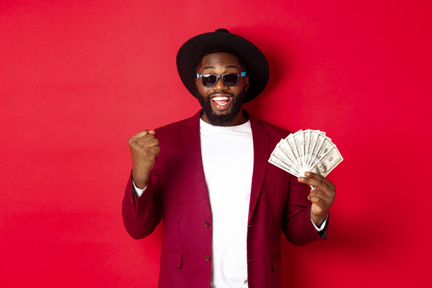 Cheerful african american guy in sunglasses, black hat and blazer, winning prize money, holding dollars and looking satisfied, standing over red background - Photo, Image