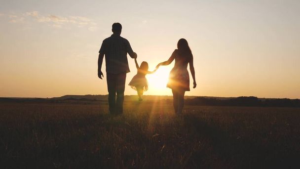 Child plays with dad and mom on field in sunset light. Family and childhood. Little daughter jumping holding hands of dad and mom in park on background of sun. Walking with a small kid in nature. - Photo, Image