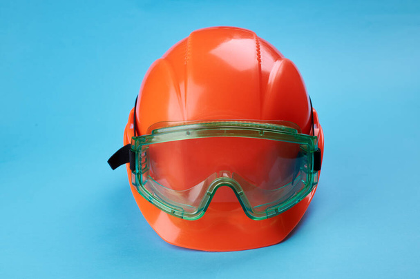 πορτοκαλί προστατευτικό κράνος και γυαλιά ασφαλείας κοντά σε αυτό σε ένα φωτεινό μπλε φόντο. προστατευτική έννοια της βιομηχανίας ενδυμάτων και κατασκευών - Φωτογραφία, εικόνα