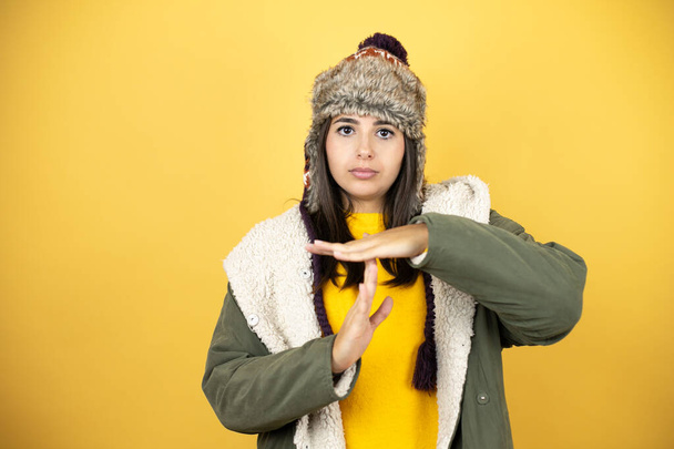 Mladá krásná žena v klobouku a zeleném zimním kabátě přes žluté pozadí odpykává si volný čas gesto s rukama, frustrovaný a vážný obličej - Fotografie, Obrázek