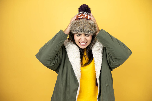 Junge schöne Frau mit Hut und grünem Wintermantel über gelbem Hintergrund leidet unter Kopfschmerzen verzweifelt und gestresst, weil Schmerzen und Migräne mit den Händen auf dem Kopf - Foto, Bild