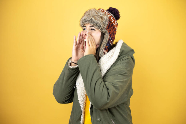 Junge schöne Frau mit Hut und grünem Wintermantel vor gelbem Hintergrund schreit und schreit laut mit den Händen auf dem Mund zur Seite - Foto, Bild