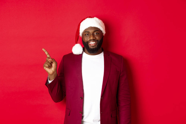 Koncepcja świąt, przyjęć i świąt. Przystojny czarny mężczyzna w czapce Mikołaja wskazując palcem w lewo, pokazując reklamę, stojąc szczęśliwy nad czerwonym tle - Zdjęcie, obraz