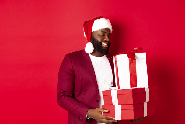 Рождество, Новый год и концепция покупок. Счастливый африканский американец выглядит тронутым и благодарным за подарки на Рождество, получая подарки, нося шапку Санты, красный фон - Фото, изображение