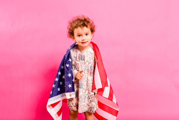 Twee jaar oud meisje poseren sympathiek met de Amerikaanse vlag op haar schouders, geïsoleerd op een roze achtergrond. - Foto, afbeelding
