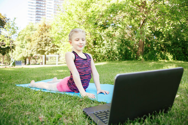 Niña caucásica haciendo ejercicio deportivo al aire libre en línea. Video yoga estirándose en Internet. Kid aprendizaje de formación en el patio trasero en casa con el ordenador portátil. Nueva normalidad. Distancia social en coronavirus. - Foto, imagen