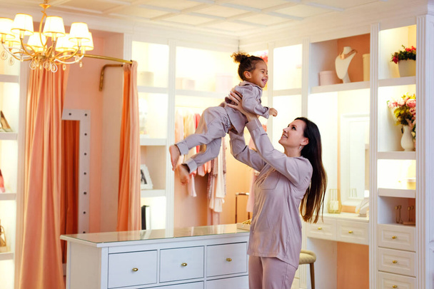 Madre caucasica e figlia afroamericana in pigiama rosa e bigodini nello spogliatoio e nella camera da letto. Madre getta la figlia tra le braccia - Foto, immagini