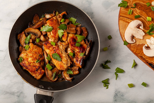 Flat lay image of a non stick frying pan with a chicken breast sautee inside. Meat is seasoned with herbs, tomato, spices, parsley, spring onions and mushrooms. There is a chopping board  - Foto, afbeelding
