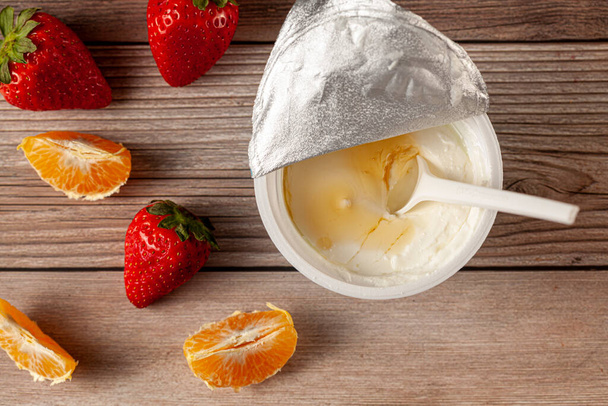 Een wit plastic kopje Griekse yoghurt met een afgeschilde aluminiumfolie afdichting en een wegwerp plastic lepel erin gedompeld geserveerd met mandarijnenschijfjes en aardbeien op hout. Gezonde snelle snack. - Foto, afbeelding