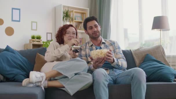 Steadicam de pelirroja chica caucásica comiendo palomitas de maíz de un tazón en manos de un tipo de raza mixta. Pareja sentada en el sofá en casa, riéndose de la película - Imágenes, Vídeo