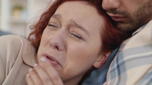 Zbliżenie młodej niebieskookiej białej kobiety z rudymi włosami płaczącej na piersi nierozpoznawalnego partnera w szachownicy trzymającego ją w ramionach - Materiał filmowy, wideo