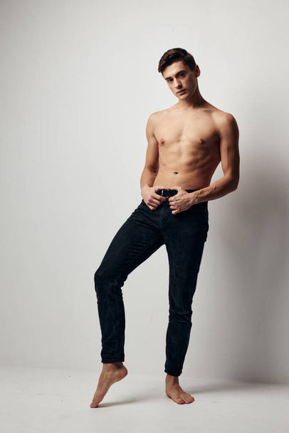 Un joven guapo en pantalones está de pie en una pierna y sostiene sus manos en su cinturón - Foto, Imagen