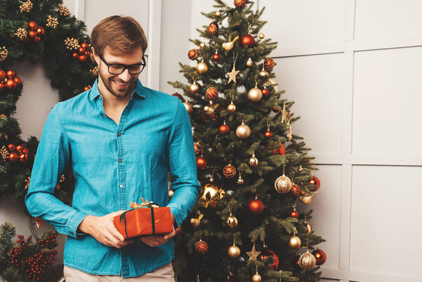 Portrait of smiling handsome man holding gift. Sexy bearded male posing near Christmas tree with present. Celebration concept. New year party. He waiting for his friends  - 写真・画像