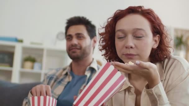 Lähikuva punatukkainen valkoihoinen tyttö ja hämärtynyt Mixed-Race mies istuu kotona, syö popcornia punainen ja valkoinen raidallinen paperi laatikot, katsella mielenkiintoinen elokuva - Materiaali, video