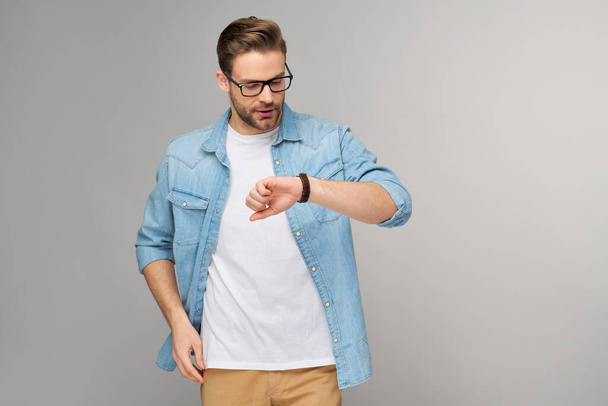 Portrait of young handsome caucasian man in jeans shirt looking at hand watch standing over light background - Фото, зображення