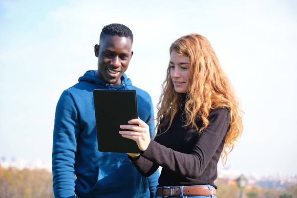 Homem negro africano e mulher caucasiana ruiva olhando para seu tablet em um parque. Jovem casal multirracial. - Foto, Imagem