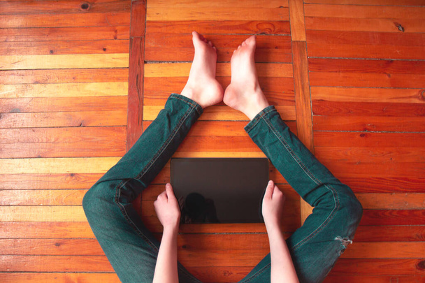 Teenager sedí na dřevěné podlaze a dívá se na obrazovku tabletu. Tableta mezi nohama. Domácí vzdělání. Selektivní soustředění. Horní pohled - Fotografie, Obrázek