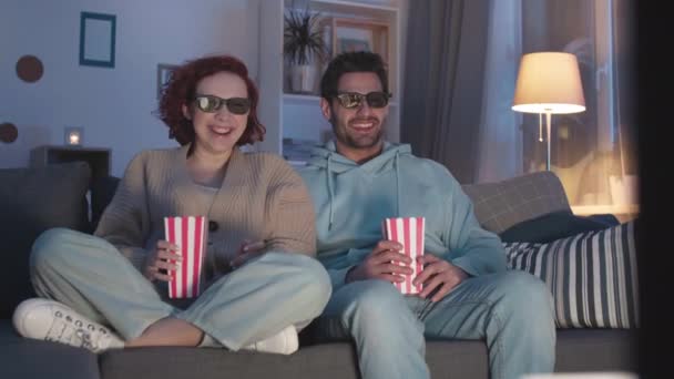 Steadicam of Mixed-Race young man sitting next to red-haired Caucasian woman in lotus pose on couch in apartment, holding popcorn boxes, wearing 3D glasses, laughing at movie - Footage, Video