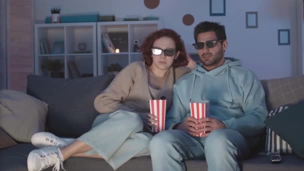 Seguimiento de la derecha de la pareja joven con gafas 3D, sentado en el sofá en la sala de estar, viendo la película, asustándose, rociando palomitas de maíz sobre sí mismos, y luego riendo - Metraje, vídeo
