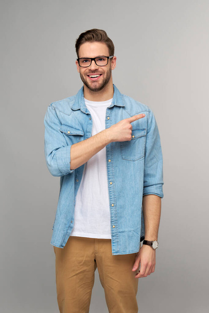 Happy young handsome man in jeans shirt pointing away standing against grey background - Foto, Imagem