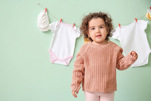 Cute baby girl w pobliżu stylowe ubrania wiszące na ścianie kolor - Zdjęcie, obraz