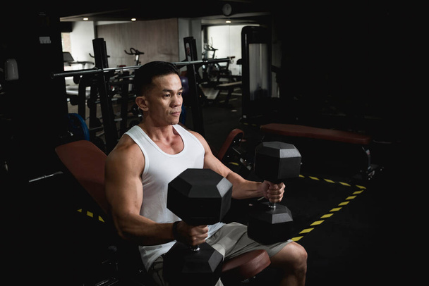 Ein athletischer asiatischer Mann hebt ein paar schwere Sechshanteln und bereitet sich mental darauf vor, Brust und Schultern zu trainieren. - Foto, Bild